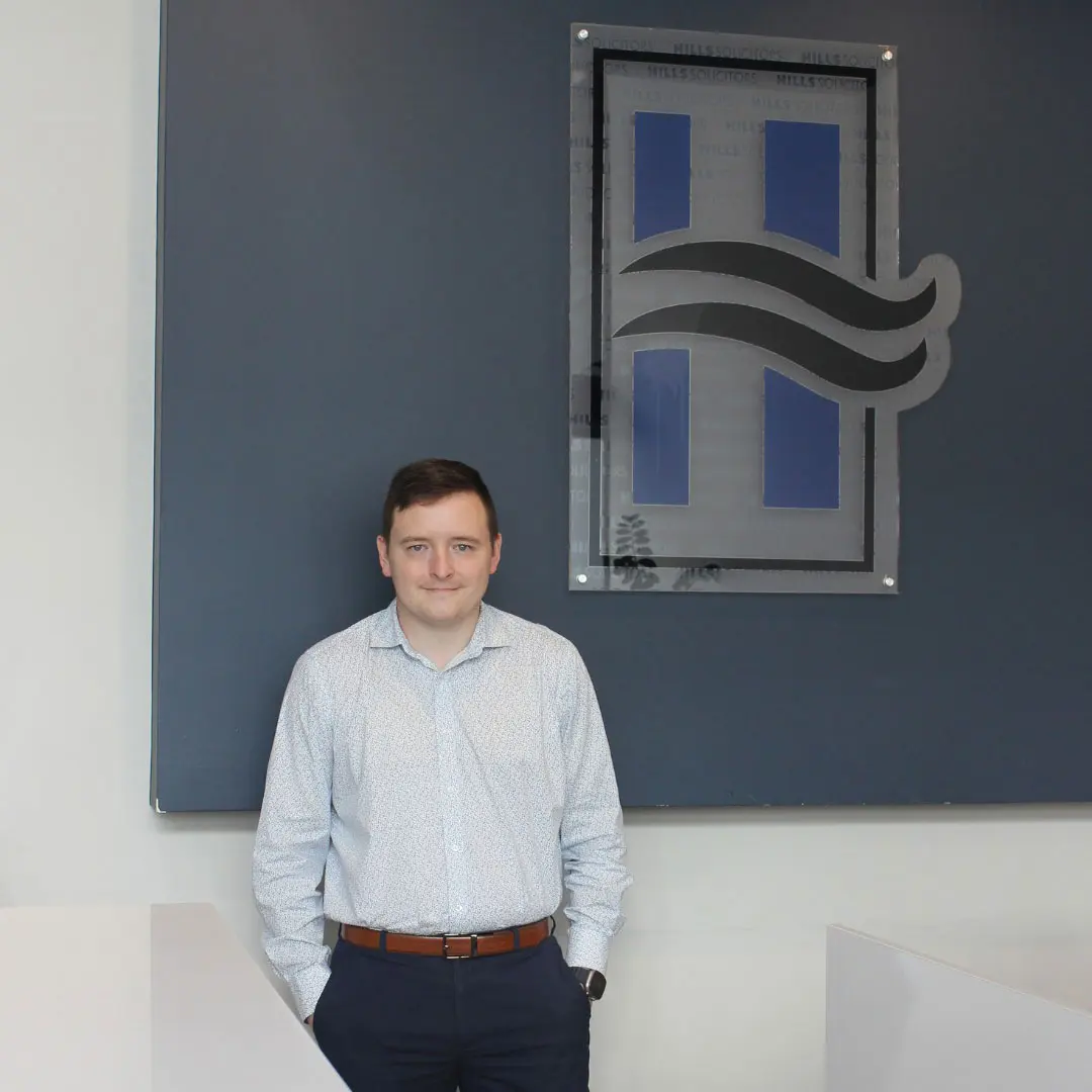 A Hills Solicitors solicitor standing in the firm's Maitland reception, beside the Hills Solicitors emblem
